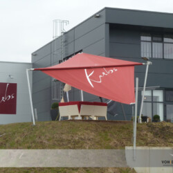 Soliday-Sonnensegel mit Werbeaufschrift Aufrollbares Sonnensegel in rot mit weißem Logo-Werbeaufdruck vor einem Gewerbebetrieb