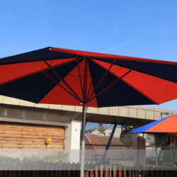 Rundschirm Blau-orange segmentiert auf Dachterrasse Runder Großschirm mit blauen und roten Tuchsegmenten auf einer Dachterrasse einer Kita